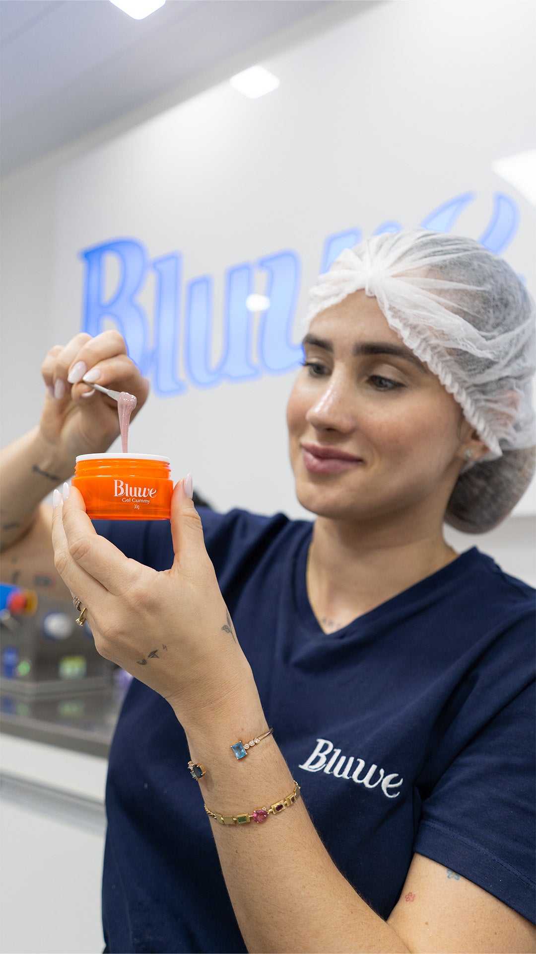 Lara Machado sorrindo e manipulando um pote com gel para unha de gel. Ela está usando touca, um uniforme azul da marca Bluwe, pulseiras e anéis.