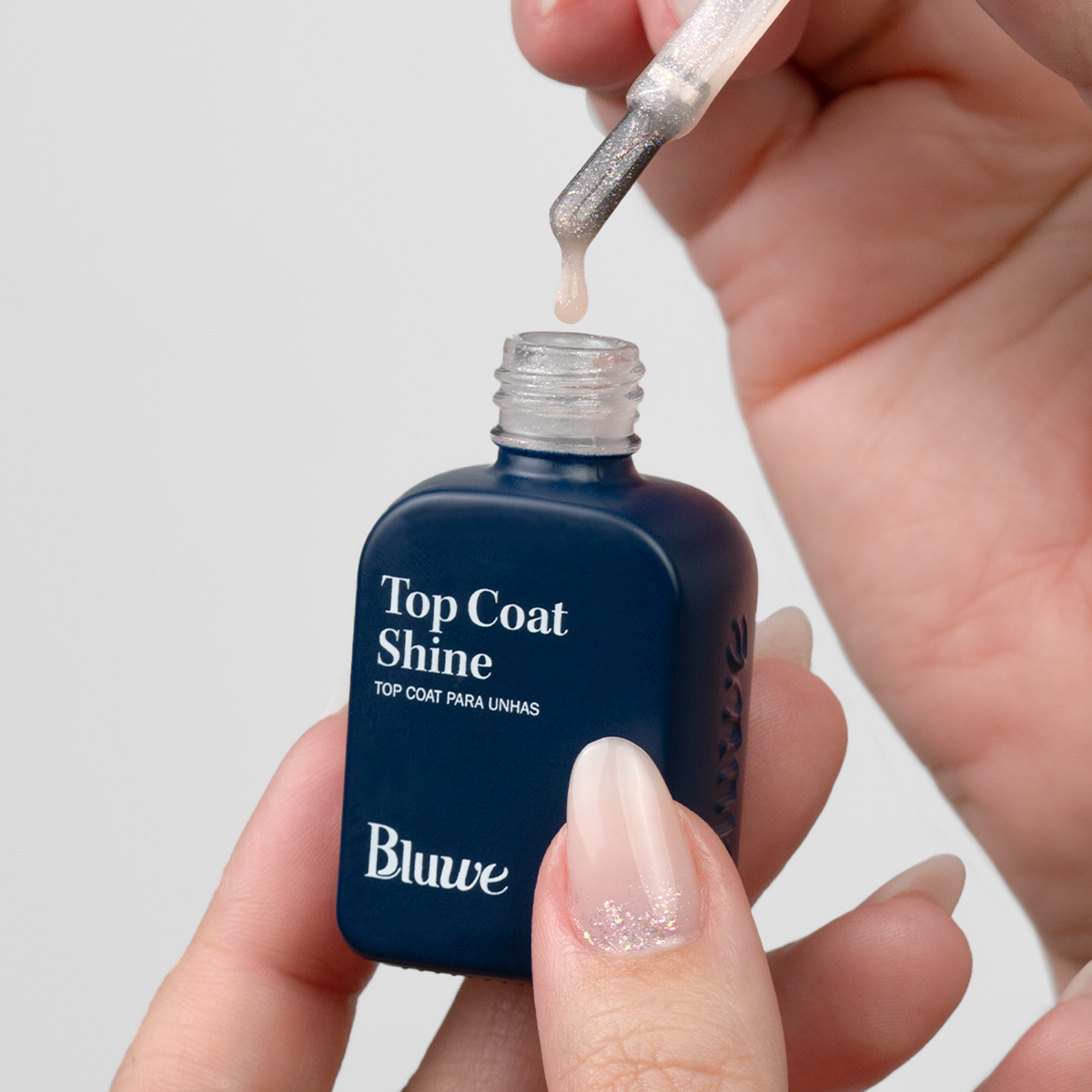 Duas mãos manipulando o produto Top Coat Shine em um vidro em formato de esmalte