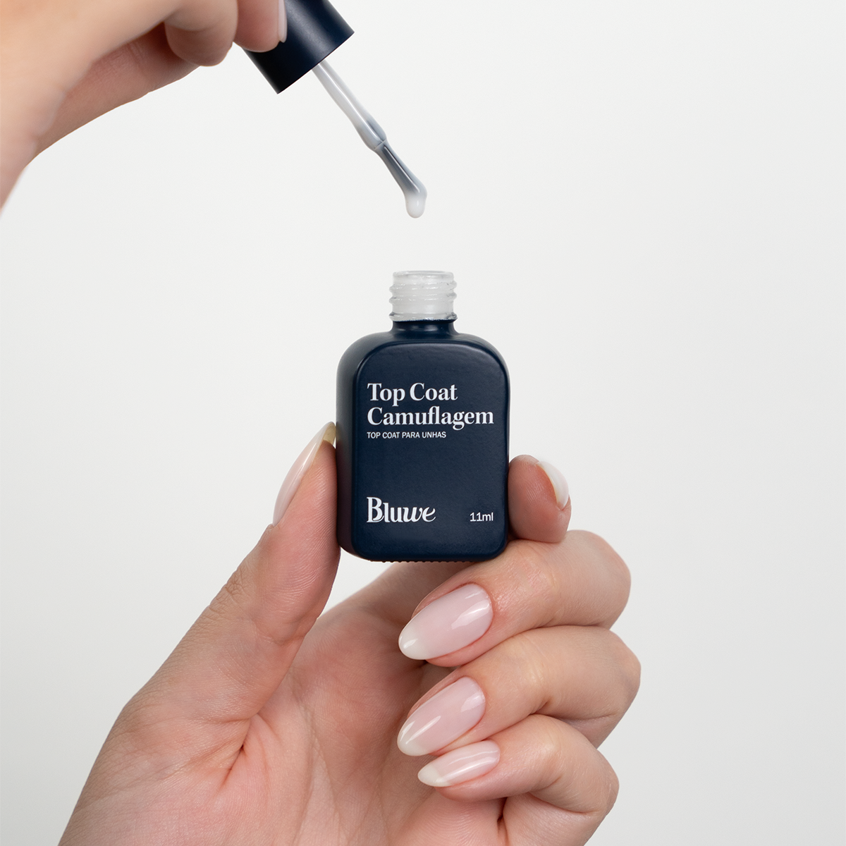 Produto Top Coat Camuflagem da marca Bluwe. Duas mãos segurando e manipulando o vidro em formato de esmalte