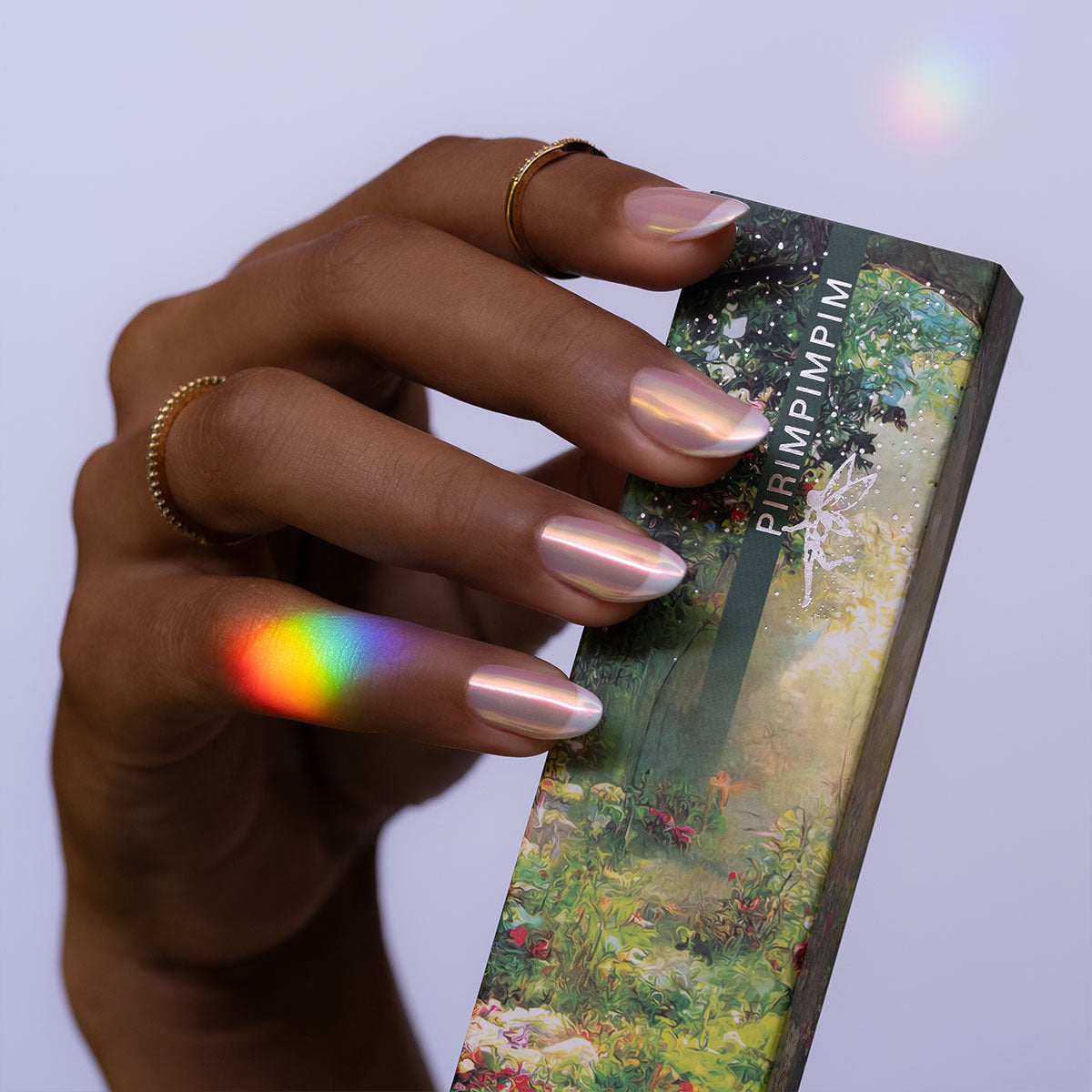 Mão da modelo segurando a caixa do kit de pós cromados, utilizados para unhas de gel, chamado Pirimpimpim